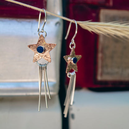 A pair of dangling earrings hanging from a dried grass stem. Each earring has a hammered gold star-shaped charm with a small dark blue stone in the center, connected to a silver-toned fishhook and long dangling bar charms.