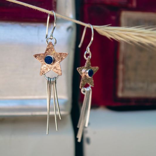 A pair of dangling earrings hanging from a dried grass stem. Each earring has a hammered gold star-shaped charm with a small dark blue stone in the center, connected to a silver-toned fishhook and long dangling bar charms.
